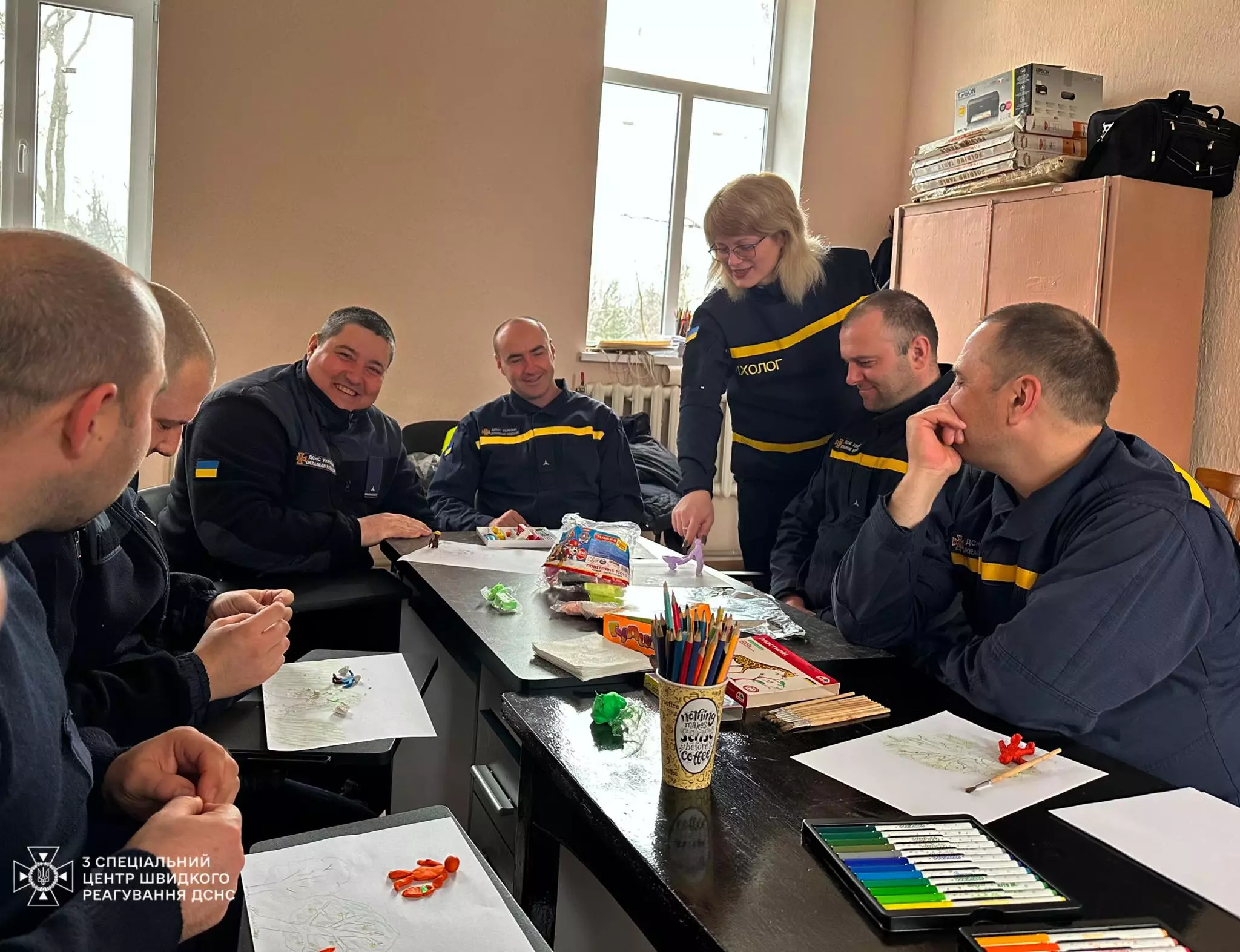 На фото зображені сапери, що сидять за столом і психологиня, яка розглядає зліплену з пластиліну фігурку