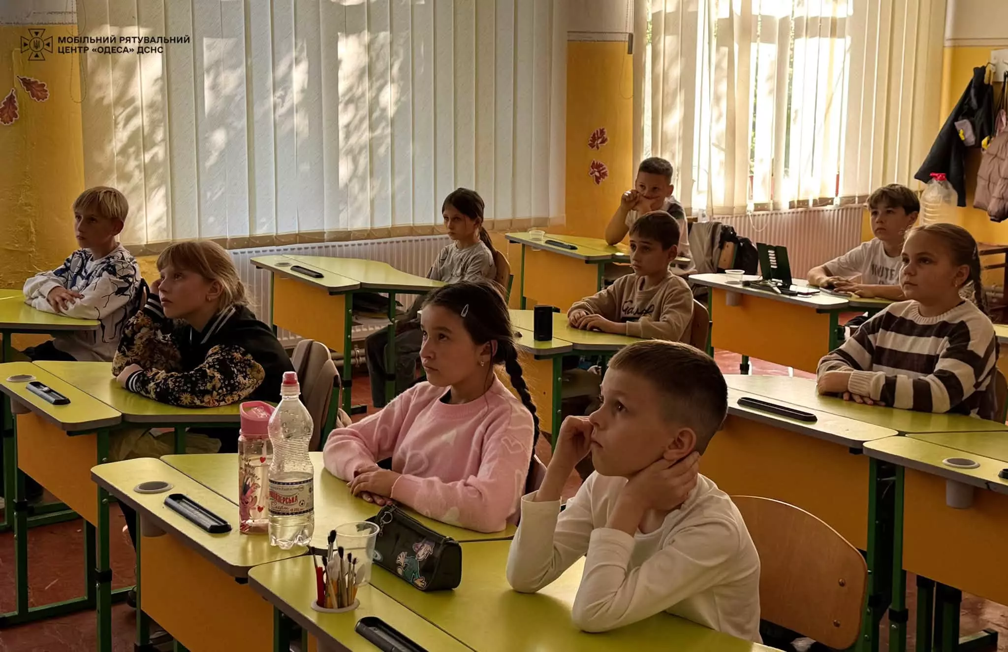 На фото діти 4 класу сидять за партами і уважно дивляться відео на телевізорі