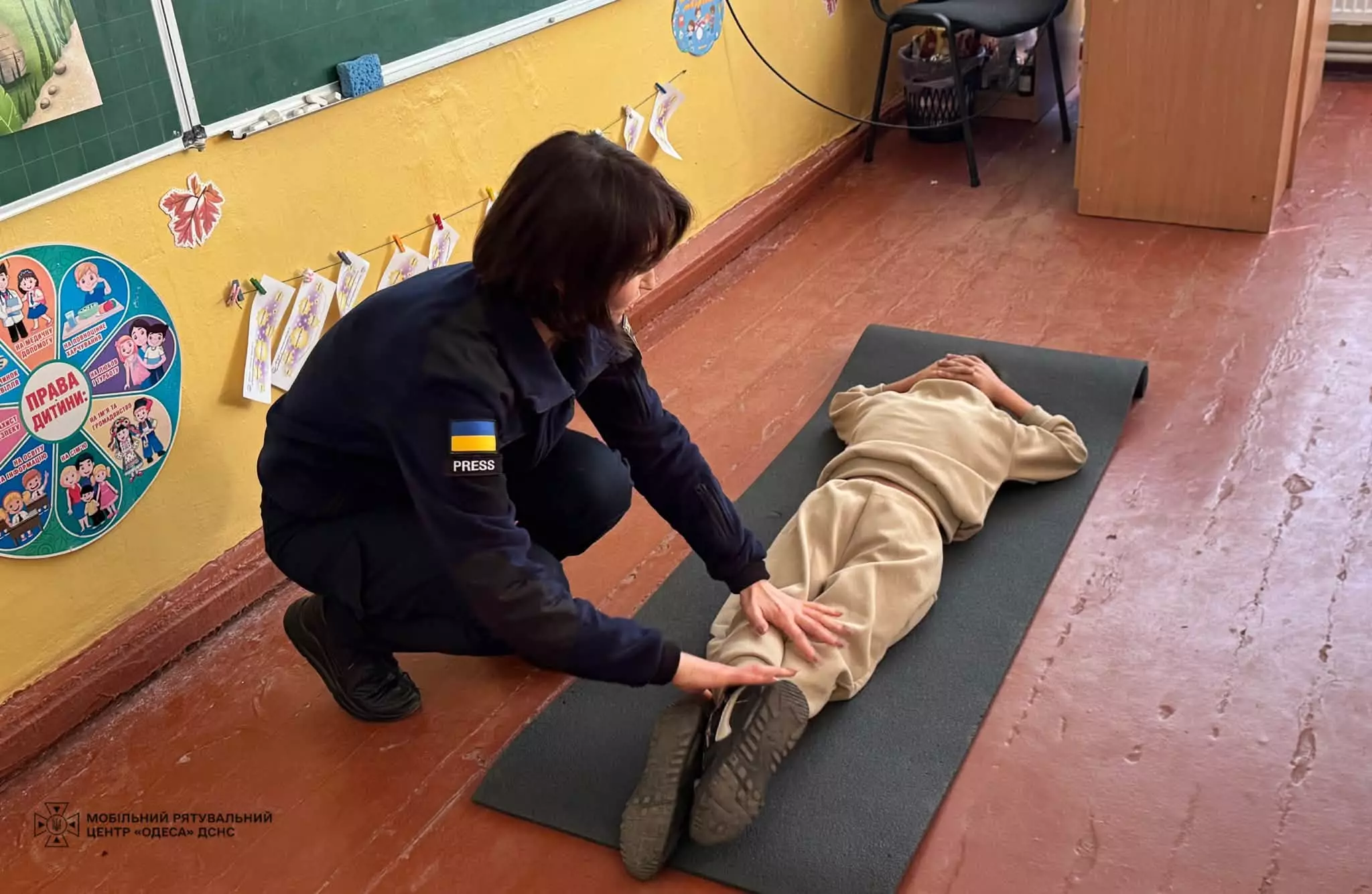 На фото рятувальниця пояснює як правильно лягти на землю, якщо почув вибух поблизу, учень лежить на килимку обличчям донизу